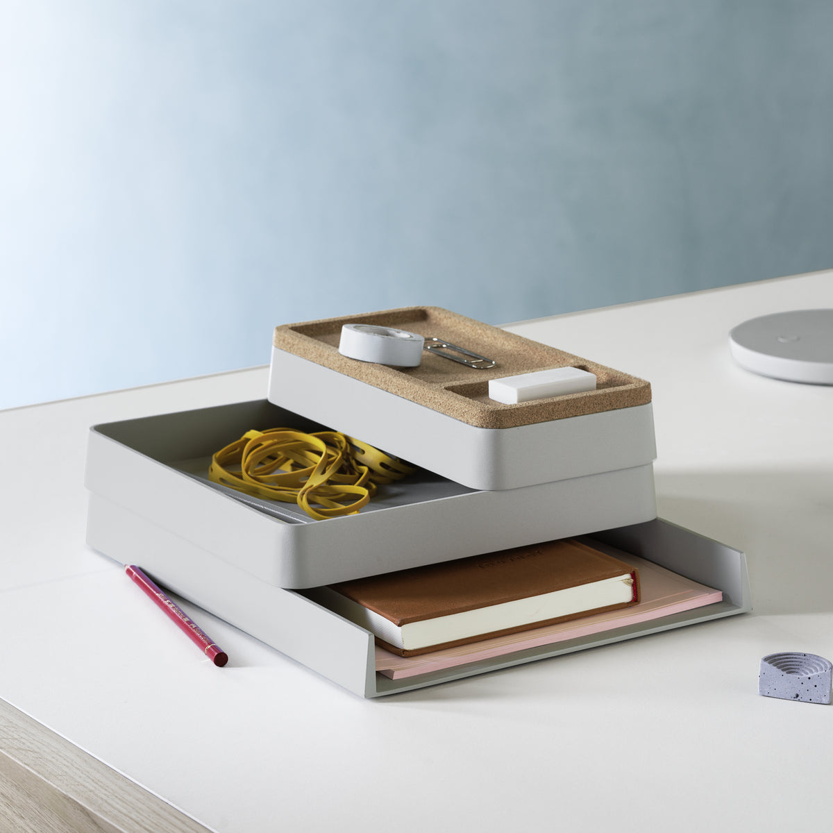 Muuto Arrange Desktop Series in grey with a cork lid, holding stationery and notebooks on a desk beside a grey lamp.