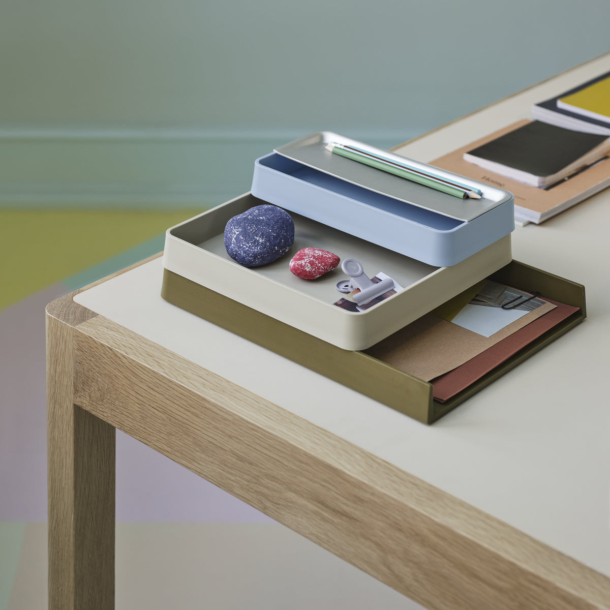 Muuto Arrange Desktop Series in olive-green, grey and light blue with a metallic lid, holding stationery on an oak-framed desk.